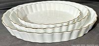 Three white porcelain oval serving trays nesting inside each other, showing wavy edges and gold trim, photographed on black background.