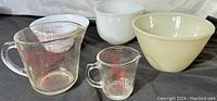 Wide view showing all five items: two milk glass mixing bowls (white and cream colored) and three clear glass measuring cups with red measurement markings.