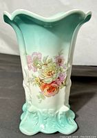 Side view showing ruffled rim, turquoise gradient, embossed floral design with pink, orange, and white roses, and ornate footed base in turquoise.
