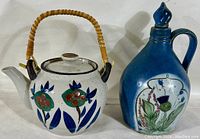 Front view of tea pot with wicker handle displaying floral design alongside Buchan thistle stoneware decanter with blue glaze and floral motif.