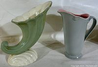 Two vintage pottery pieces side by side on white fabric background: a green and white cornucopia vase and a pale blue pitcher.