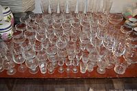 Top-down view of assorted clear glassware arranged on a wooden table including wine glasses, shooters, champagne flutes, snifters, and branded Olympic glasses.