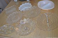 Photo showing assorted clear glass serving dishes with metal handles and patterned edges alongside white and beige ceramic dishes on wooden table.