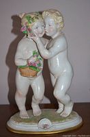 Front view of porcelain cherub figurine showing two nude children with basket of flowers on grass base.