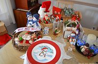 Wide view of Christmas basket items on table showing plates with snowman design, figurines including Santa and snowmen, ornaments, candles, and garland
