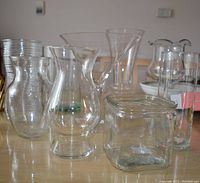 Multiple clear glass vases of different shapes and sizes grouped together on a wooden table. Includes tall, bulbous, square, and flared rim styles.