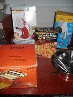 Photo of kitchen gadgets boxed and loose items on surface: pasta maker boxes, cookie press box, food mill, and grater.