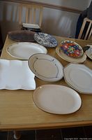 Assortment of seven serving platters arranged on a wooden table showing variety of shapes and patterns including floral designs and white ceramic pieces. Two platters with visible chips.