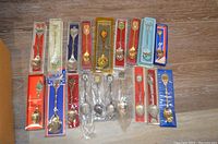 Photo shows nineteen souvenir spoons arranged on a wooden surface, each in its original packaging made of plastic or cardboard. Packaging colors include red, blue, gold, orange, and clear plastics. Spoon designs vary including floral motifs, regional emblems, and colorful enamel effects.