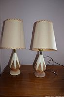 Front view of the two vintage table lamps side by side showing their beige and brown ceramic bases and matching original beige fabric shades with braided trim.