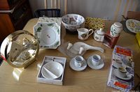 Photo showing various ceramics on a table including plates, bowls, figurines, cups, creamer and sugar set, boxed white dish, espresso coffee sets, and a gold tone clock.