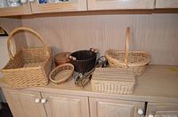 Wide view of seven wicker baskets of different sizes and styles placed on a light wood cabinet, primarily light brown wicker with some black and colored accents
