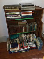 Wooden bookshelf with three shelves holding hardcover books and a lower stack of paperbacks