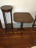 Two wooden tables side by side: a tall round plant stand or lamp table with lower shelf on the left, and a square tilt top table with turned pedestal base on the right, placed against a white wall on wood flooring.
