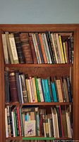Full bookshelf showing three shelves filled with various hard and soft cover books of different sizes and colors.