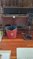 Front view of wood fireplace bin with metal liner, fireplace tools on stand, and small decorative red metal bucket in front.
