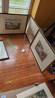 Group view of three framed paintings on hardwood floor near window