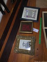 Three framed pieces on wooden surface: ornate gilded frame at bottom, small plaque frame center, black frame print at top right.