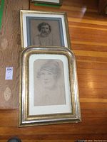 Two framed older portrait pencil sketches laid on wooden floor, one larger portrait of a man with curled hair, one smaller portrait of a woman with hat and earring.