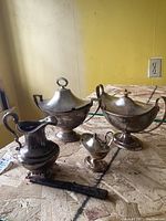 View of sterling creamer, two silver-plated sauce tureens, small salt cellar, and cutter in scabbard on plywood surface.