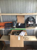 Wide view showing all items on metal shelves: two socket sets, cordless jigsaw, mouse sander, and cardboard box containing miscellaneous tools and instructions.
