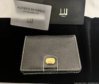 Closed black Alfred Dunhill leather card holder with gold-tone logo plaque on front, authenticity cards and black box behind.