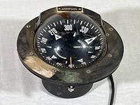 Front angled view of Ritchie Powerdamp gimbal mount compass showing black dial with numeric degree markings and directional arrows, mounted in metal frame with visible mounting screws and wiring.