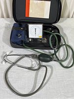 Full view of open black leather zippered case containing blood pressure monitor with green rubber tubing, blue cuff, and attached stethoscope. Instruction papers are visible inside the lid.