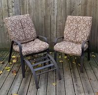 Two patio chairs with brown brushed-iron frames and rubber slats, one reclined with footrest and both fitted with patterned cushions, on wood deck against fence.