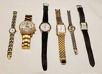Six wrist watches laid flat showing variations in band styles and colors including metal link bands in gold and silver tones and one black leather band. Watches vary in dial shapes and sizes.