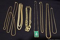 Seven gold-colour chain necklaces laid flat on dark surface showing variety of chain styles and lengths, including one with a green rectangular pendant with white Asian character.