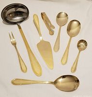 Full set of eight gold-coloured stainless steel Solingen serving utensils arranged on a white cloth, including ladle, fork, pie server, tongs, and spoons.