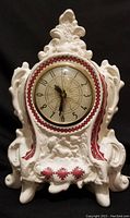 Front view of ceramic clock showing ornate moldings, clock face and red and black embroidery pattern.