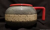 Side view of the curling rock shaped ceramic mug showing speckled gray and beige bands with a red handle and rim.