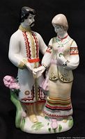 Front view of the ceramic figurine showing detailed handpainted traditional clothing on the man and woman, floral base with pink flowers.