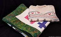 Folded quilt with green leafy border and multicolor Chop Suey blocks alongside off-white hand-knitted sweater with patterned yoke, on black background.