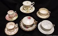 Full view of all six varied floral china teacups with matching saucers on black background