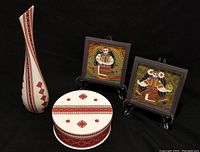 Two framed textured paintings of women in Ukrainian folk costumes, a tall ceramic vase, and matching round lidded dish with red geometric patterns on white background, all displayed on black.