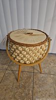 Photo of the mid century round sewing basket showing woven rattan body with pleated fabric lid and wooden tripod legs.