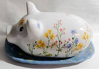Side view of the white ceramic pig butter dish lid decorated with small floral details on a blue floral base plate.