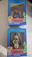 Photo showing two boxed Brass Key Collection Rudolph The Red-Nosed Reindeer hand-crafted glass ornaments with North Pole sign and tree graphics on the front of the boxes.