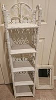 Full view of the tall vintage white wicker shelf with five shelves and decorative curved top, shown next to the small rectangular wicker framed mirror.