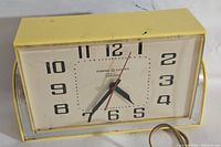 Yellow General Electric 2H47 electric clock front view showing clock face with black numbers, black hour and minute hands, and a red second hand. Clock is rectangular with yellow casing and silver metal accents on sides.