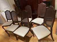 Seven wooden dining chairs shown grouped together, displaying cane backs and white upholstered seats on curved legs.