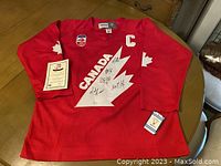 Full view of red Team Canada jersey laid flat showing overall condition, signatures and certificate at side