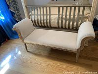 Full side view of the fabric bench showing scroll armrests, carved wooden legs and frame, light beige upholstery.