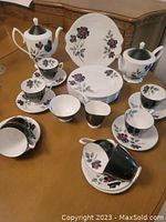 Front and top view of the Royal Albert Masquerade tea set for 8 on a wooden table, showing teapot, sugar bowl, creamer, 8 plates, cups and saucers, 2 small bowls, all with floral pattern and black accents.