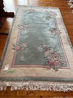 Full view of rectangular area rug with floral border design placed on wooden floor, showing pale blue and off-white colors with pink flower clusters and fringe on one end.