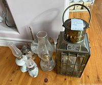Photo showing a group of assorted lamps including a large brass lantern and smaller kerosene lamps with glass chimneys on a wooden surface
