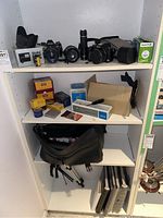 Wide view of shelves showing cameras, lenses, film rolls, camera bags, a tripod, and photo albums on shelf.